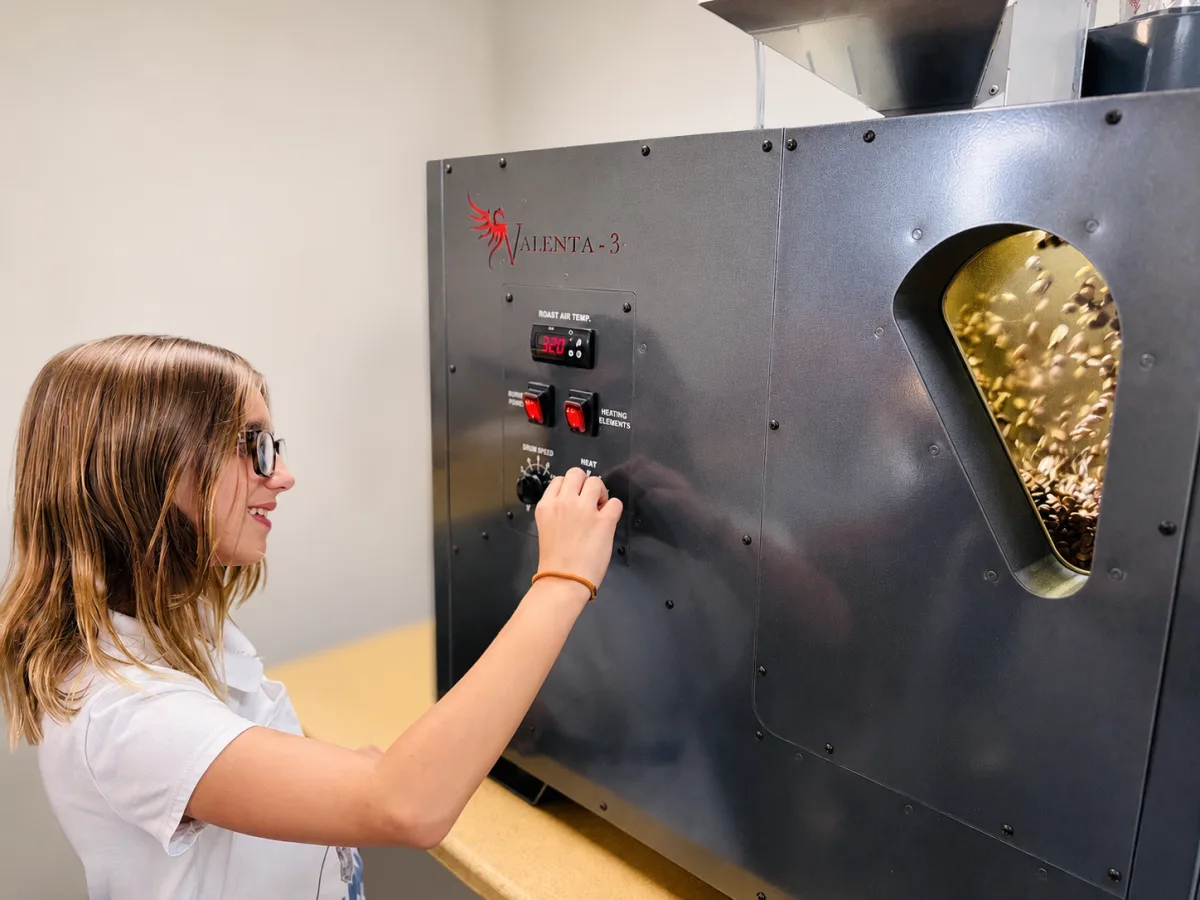 A founder at the roaster controls, green beans tumbling behind the drum window.